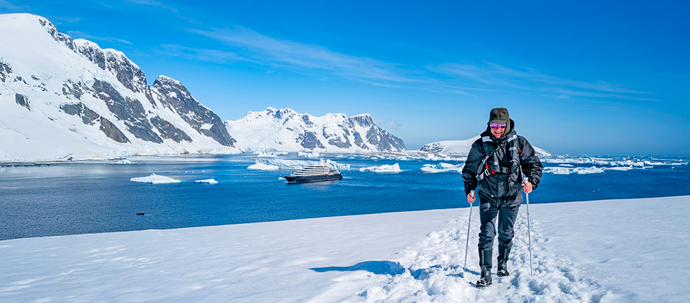 Man walking through snow in Antarctica with Scenic yacht at sea and snowy mountains in the background.