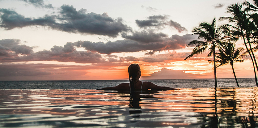 Four people paddling in an outrigger canoe off the coast of Maui with a hotel and palm trees on shore in the background.