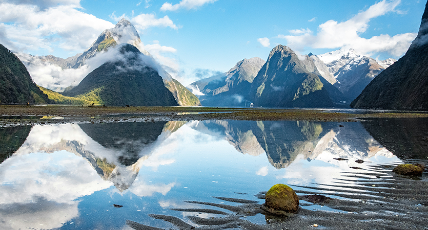 Milford Sound in New Zealand. Tall mountain peaks reflect in a body of water on a sunny day.