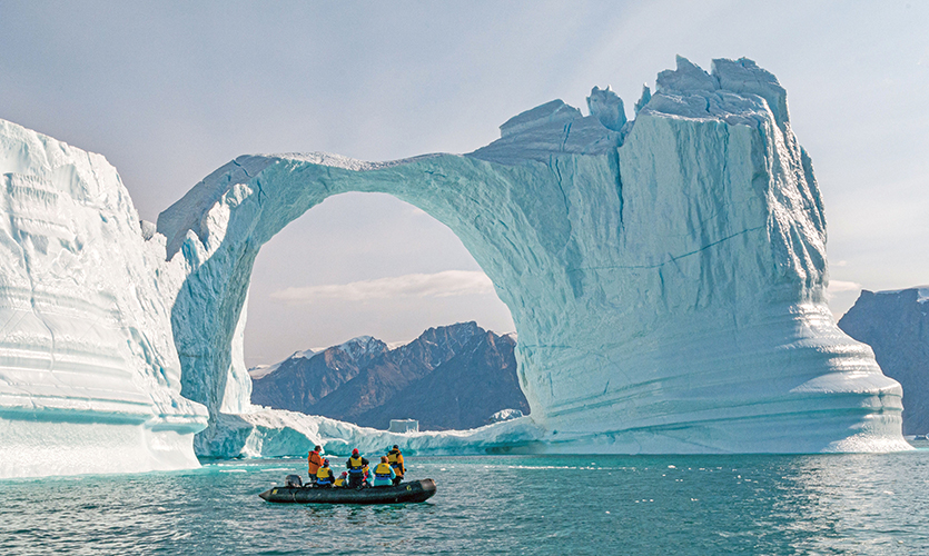 Group of people in a Zodiac boat in front of a towering iceberg in Greenland.